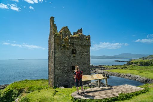 A tourist with a red backpack explores the ruins of Gylen Castle by the sea on a sunny day in Scotland