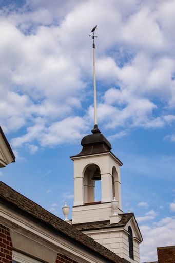 Historic bell tower with weather vane against a bright blue sky and scattered clouds, showcasing classic architecture.