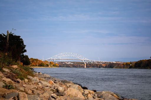 Scenic view of the Bourne Bridge over Cape Cod Canal with rocky shoreline and autumn trees under a clear sky.