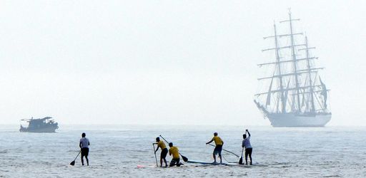 People paddleboarding near a tall ship on a misty sea, creating a serene maritime scene.