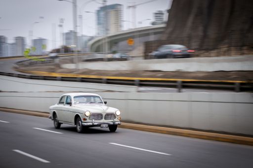 A classic vintage car driving on an urban highway with blurred cityscape in the background.