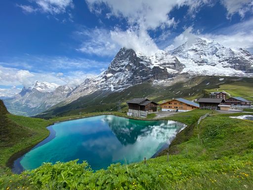 A beautiful pond surrounded by green landscapes and  wooden houses on alpine mountains background
