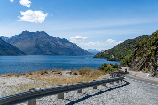 Scenic view of a winding road by a tranquil lake and majestic mountains under a clear blue sky in New Zealand.