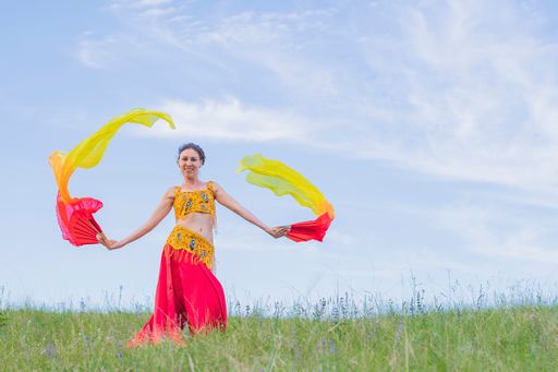Young woman in bright national dress and fiery fans in her hands is dancing oriental dance against of blue cloudy sky. Stands facing the camera.