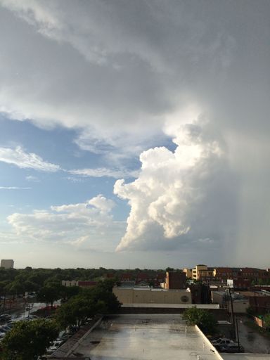 Dramatic cloud formation over a cityscape with buildings and lush greenery, highlighting the contrast between storm clouds and clear blue sky.