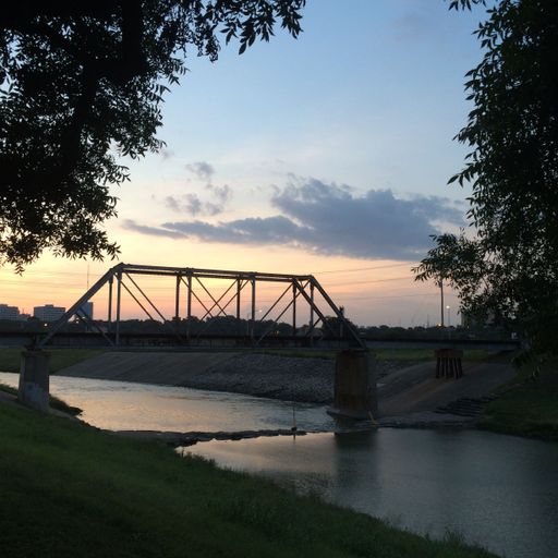 Scenic view of a bridge over a tranquil river at sunset, framed by trees, with city skyline in the background.