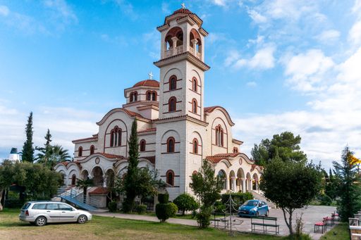 The beautiful Greek Orthodox Church of Apostle Paul and Saint Asti under a partly cloudy sky, surrounded by greenery and parked cars.