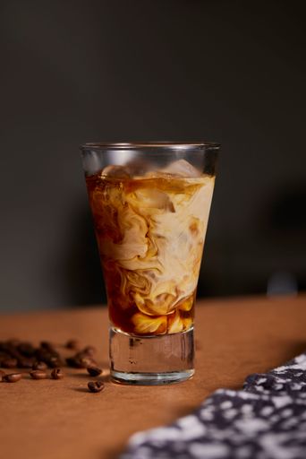 A glass of iced coffee with milk swirling, placed on a brown surface with scattered coffee beans and a patterned cloth.