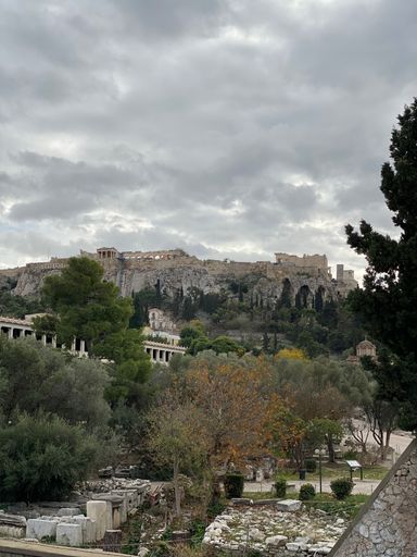 Scenic view of the Acropolis in Athens, Greece, under a cloudy sky with lush greenery and ancient ruins in the foreground.