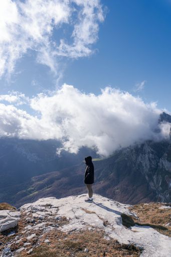 A person standing on a mountain peak, overlooking a vast valley under a bright blue sky with scattered clouds.