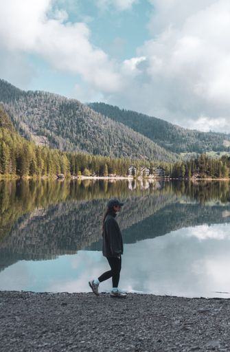 Young woman walking by a serene lake with mountain reflections under a cloudy sky, showcasing natural beauty and tranquility.