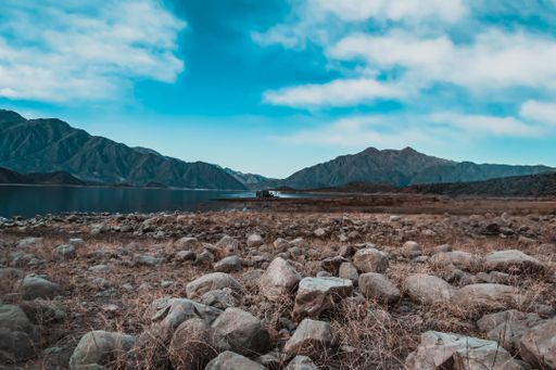A mesmerizing view of a rocky field, calm lake with hills on the background under a cloudy sky