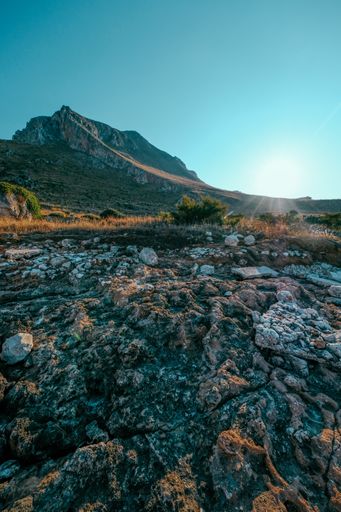 A vertical shot of rocks near a dry grassy field with mountain and a clear blue sky in the background