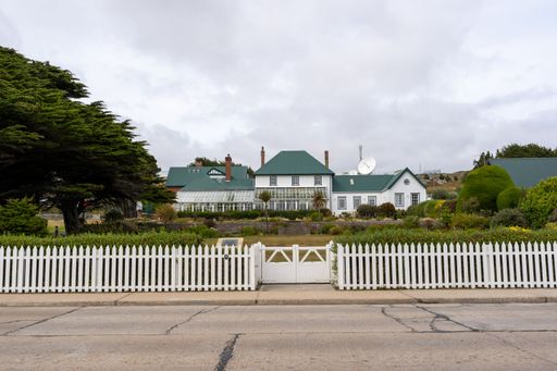 Charming colonial-style house with green roofs and white picket fence in a peaceful suburban setting under a cloudy sky.