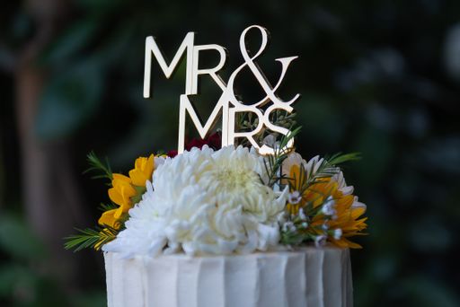 Elegant wedding cake topped with Mr & Mrs sign and fresh flowers, set against a blurred garden background.