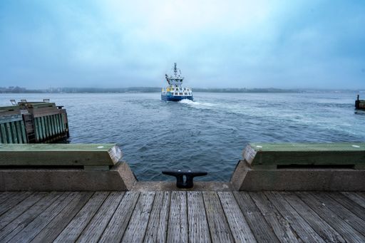 The ferry departs from the wooden pier on a misty morning, with a cloudy sky over a calm sea, creating a serene maritime scene.