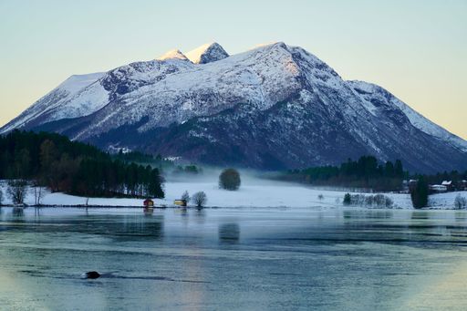 Scenic winter landscape of snow-capped mountains reflecting on a calm lake, with a small cabin nestled among trees, under a clear sky.