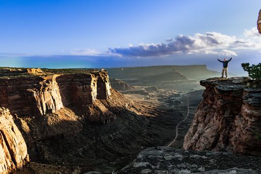 Man standing triumphantly on a cliff edge overlooking a vast canyon under a clear blue sky, capturing the essence of adventure and nature.