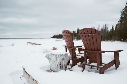 Two Adirondack chairs in a snowy landscape by a frozen lake, surrounded by trees under a cloudy sky, evoking a peaceful and serene winter scene.