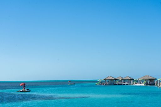Tropical beach scene with turquoise waters, overwater bungalows, and a jet ski with a red umbrella under a clear blue sky.