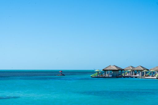 Calm sea with bungalows on stilts and a red umbrella floating object under a clear blue sky.
