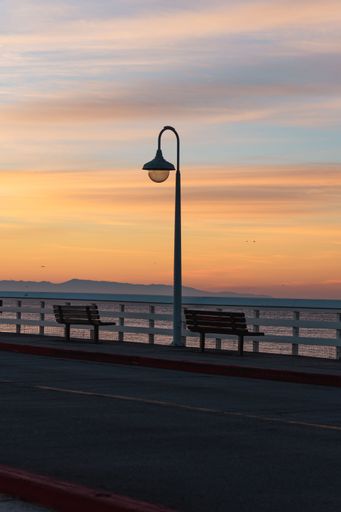 A serene seaside pier during sunset, featuring a lamp post and benches with a colorful sky in the background.