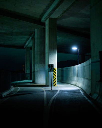 Moody night scene under a highway overpass, illuminated by a single streetlight casting shadows on the curved road.