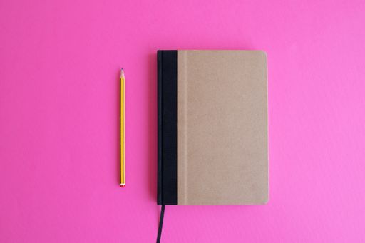 A top view shot of a beige planner and a yellow penci on a pink background