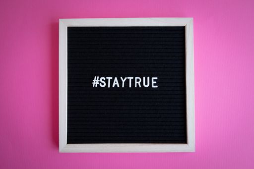 A top view shot of a blackboard with a white frame with a "#staytrue" hashtag on a pink background