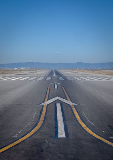 A long airport runway with a clear blue sky and distant mountains, featuring an airplane taking off in the background.