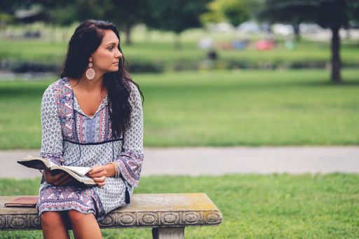 A female taking a pause from reading a bible at a park with grass and trees