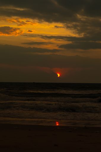 A vertical image of the setting sun partially hidden behind clouds over the ocean with its reflection on the beach in Binh Thuan, Vietnam