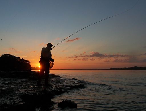 Silhouette of a fisherman casting a line at sunset by the sea, with a vibrant sky and calm waters.