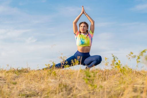 Young beautiful girl in bright multicolored tracksuit performs physical exercises to stretch muscles of legs outdoors on background of blue summer sky