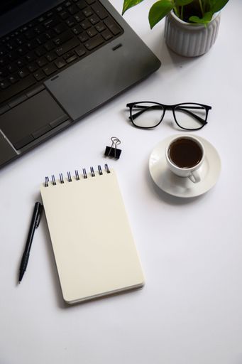 Office workspace with a laptop, notebook, pen, glasses, coffee cup, and plant on a white desk.
