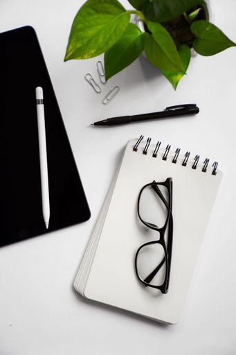 Flat lay of a workspace featuring a tablet, stylus, notebook, glasses, pen, paper clips, and a potted plant on a white background.