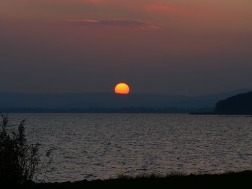 Serene sunset over a tranquil lake with a vibrant orange sun dipping below the horizon, creating a peaceful evening scene.