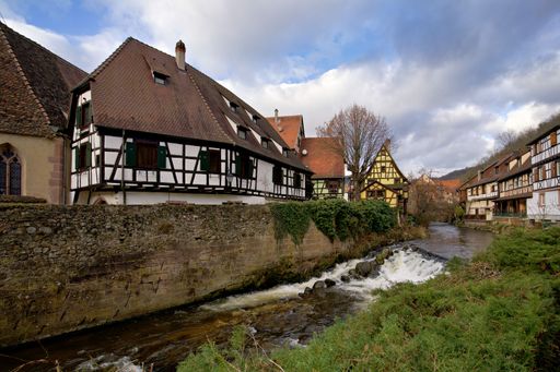 Charming Alsatian village scene with traditional half-timbered houses along a flowing river under a partly cloudy sky.