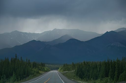 A scenic road leading through lush green forests with majestic mountains under a cloudy sky, capturing the essence of a tranquil wilderness.
