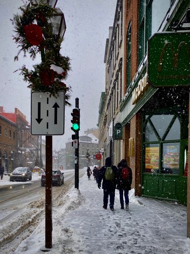 Winter street scene with pedestrians walking on a snowy sidewalk next to a McDonald's, in a city setting during snowfall.