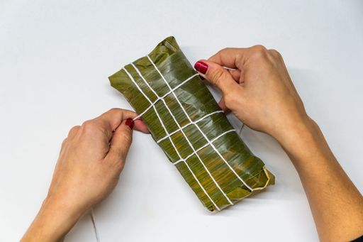 Close-up of hands tying a traditional Venezuelan hallaca with string on a white table. The image captures the essence of holiday preparation process.