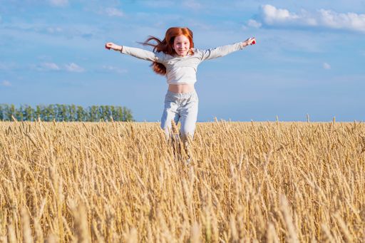 A young red-haired girl flies in a jump over a gold wheat field against a blue summer sky. Looks into the camera.