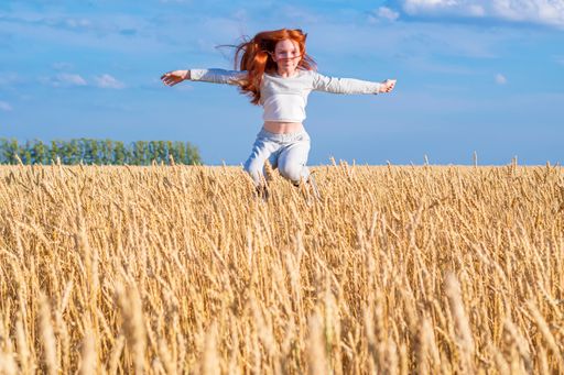 A young red-haired girl flies in a jump over a gold wheat field against a blue summer sky. Looks into the camera.