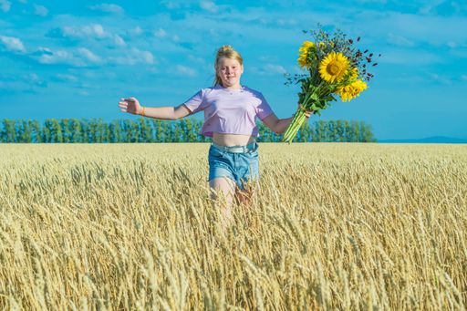 Young girl runs across golden wheat field against blue summer sky. In his hand holds bouquet of sunflowers and wildflowers. Looks into the camera
