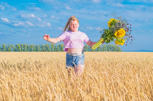 Young girl runs across golden wheat field against blue summer sky. In his hand holds bouquet of sunflowers and wildflowers. Looks into the camer