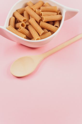 A vertical shot of a bowl of uncooked pasta and a wooden spoon on a pink surface