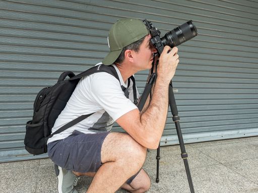 Freelance street photographer kneeling with tripod and camera, shooting urban architecture under the summer sun.