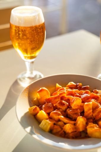 Close-up of a glass of beer and a plate of patatas bravas in soft sunlight, creating a warm and inviting dining atmosphere.