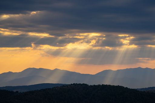 Sun rays breaking through clouds over a mountain range during sunset, casting a serene glow on the landscape.