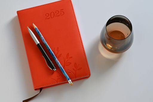 Orange 2025 planner with pens and a glass on a white table, symbolizing organization and planning.